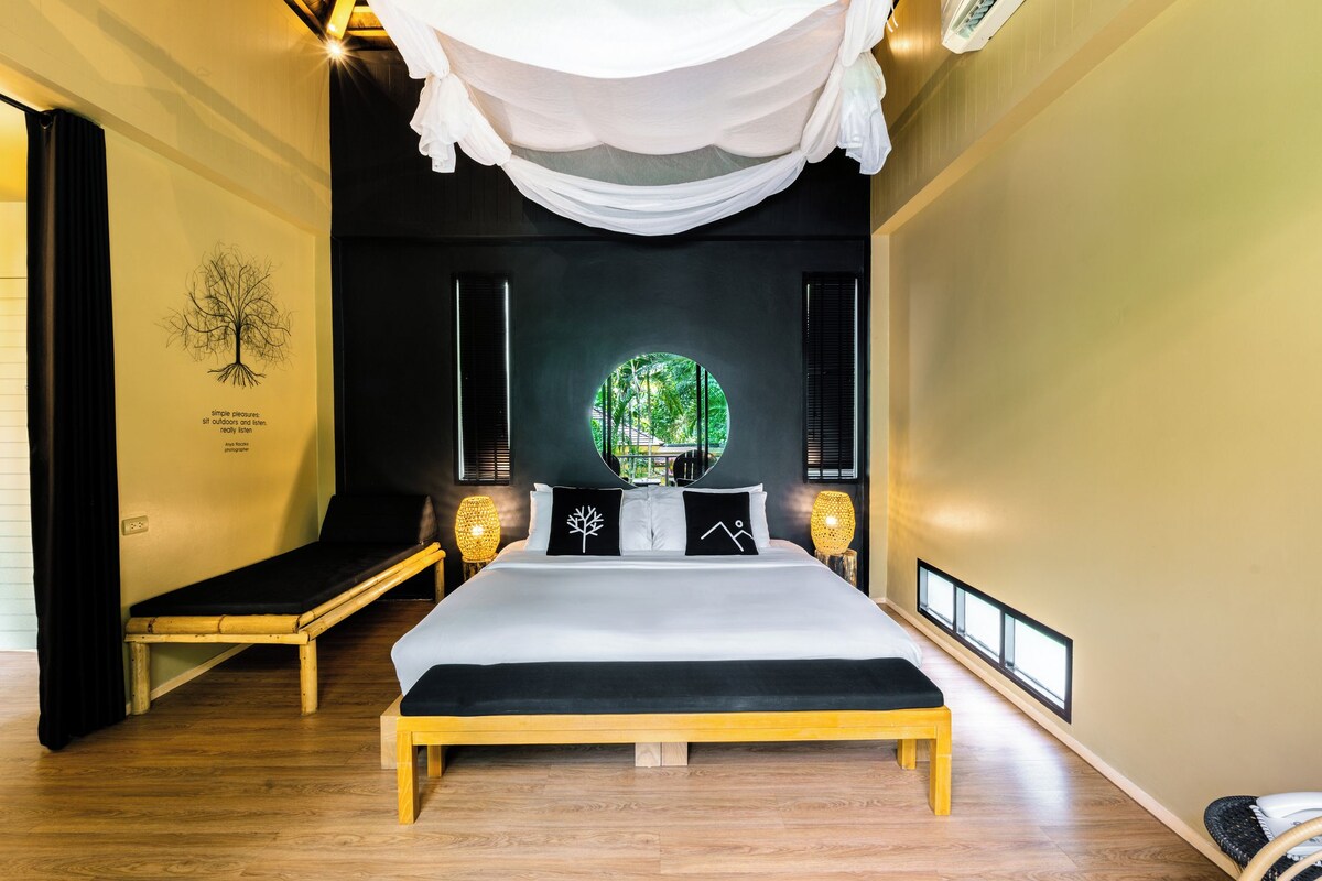 Une chambre d’hôtel avec un grand lit, un banc, une lampe fixée au mur et une fenêtre donnant sur un paysage verdoyant.