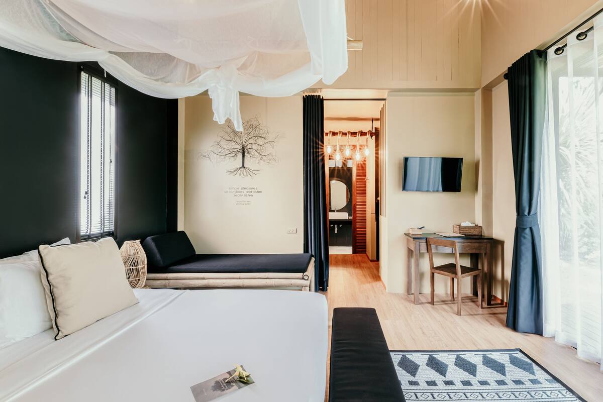 Un dormitorio con cama, mesita de noche y escritorio con silla. Hay un televisor en la pared y una ventana con cortinas.