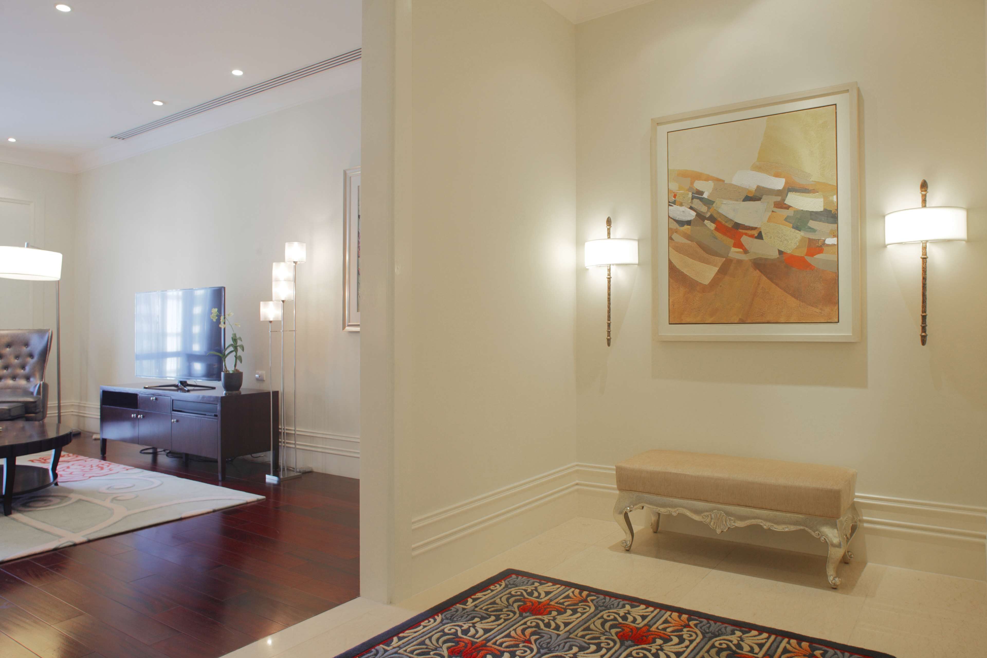 A hotel lobby with a wooden floor, a bench, a desk with a chair, a painting on the wall, and wall-mounted lamps.