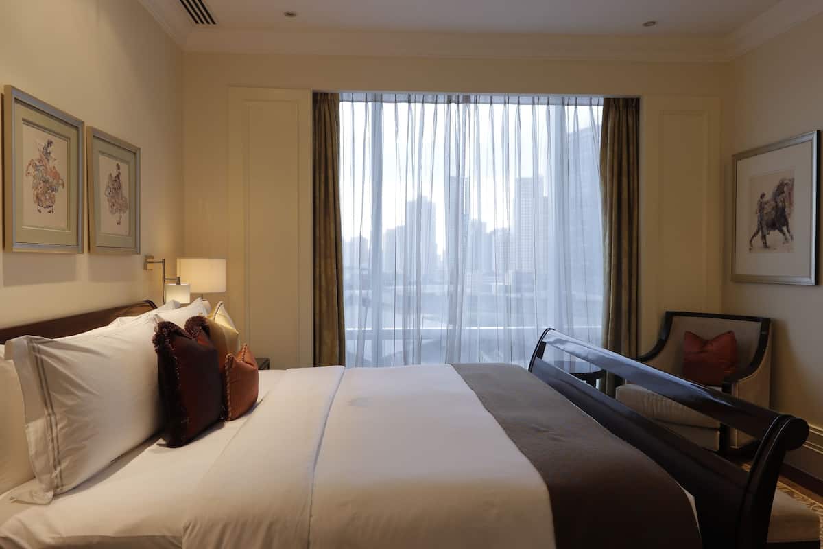 Une chambre d’hôtel avec un grand lit, une vue sur la ville à travers de fines rideaux, et des œuvres d’art accrochées aux murs.