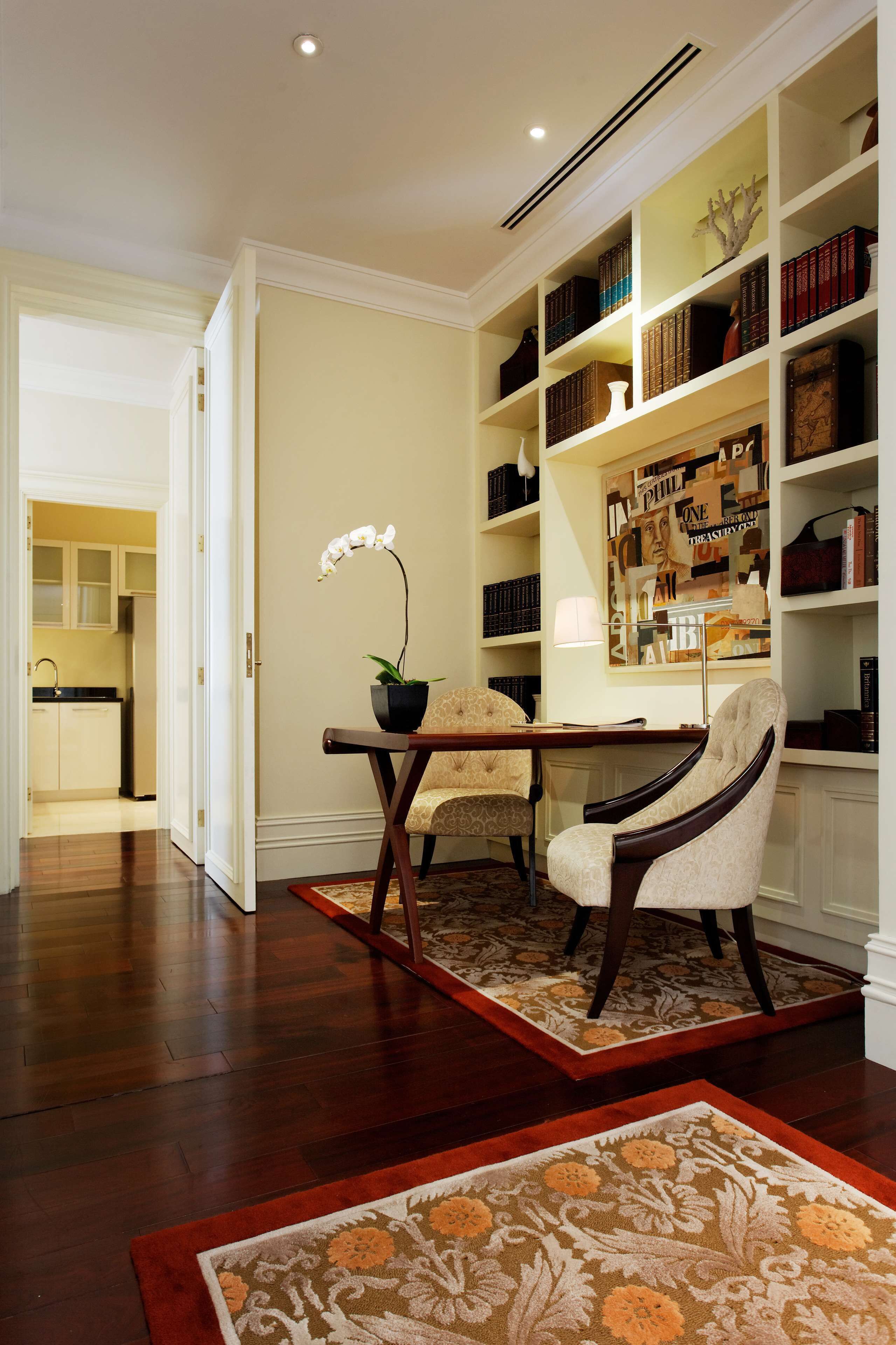 A room with a wooden floor, a desk with a chair, a bookshelf, and a potted orchid.