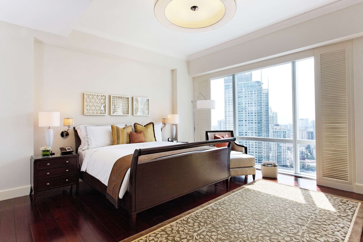 Une chambre spacieuse avec un grand lit, une table de chevet, une lampe de chevet, un lampadaire et une vue sur la ville grâce à de grandes fenêtres.