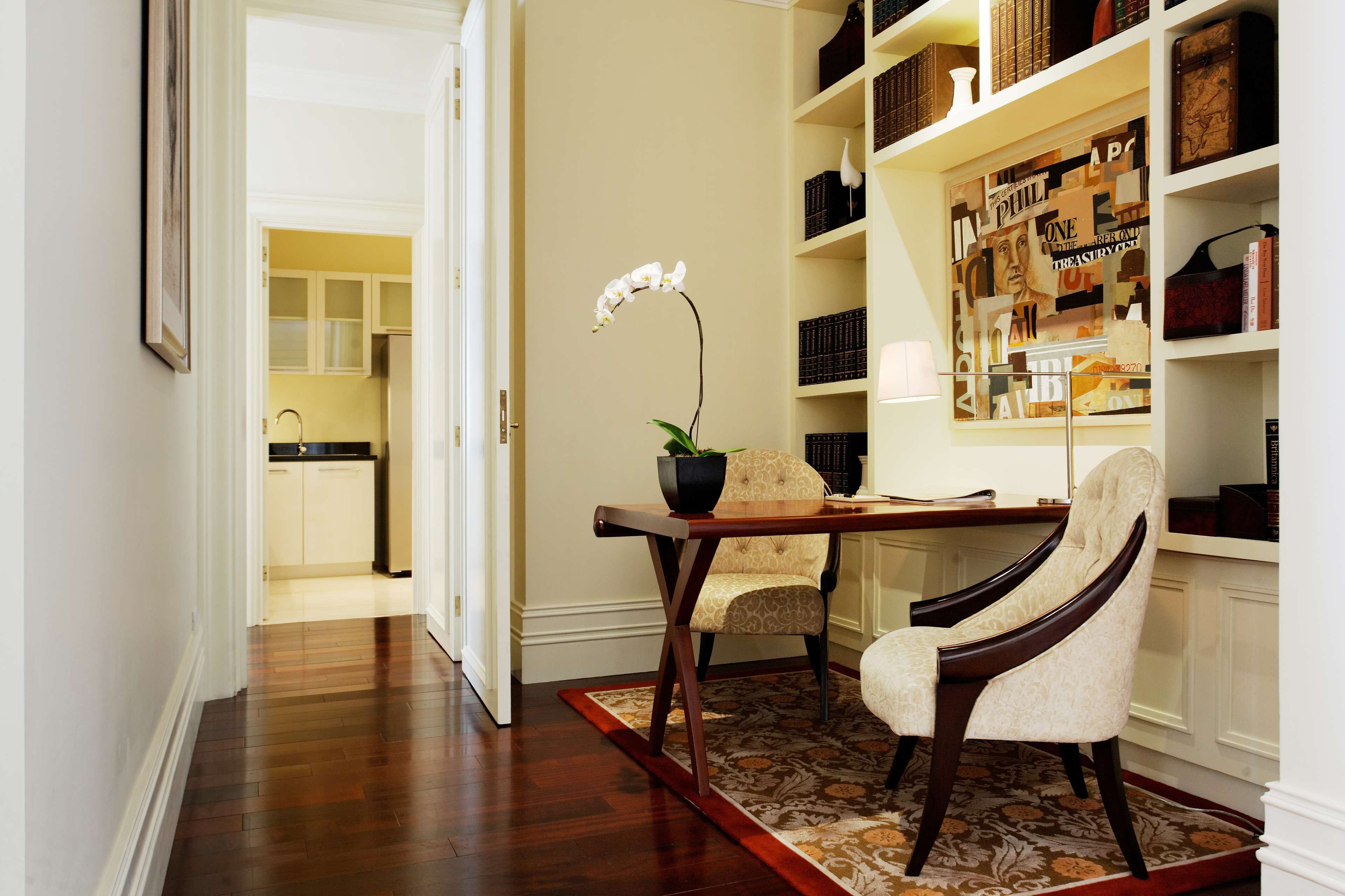 Una habitación con piso de madera, una mesa de comedor con sillas, una orquídea en maceta y una estantería llena de libros y adornos.