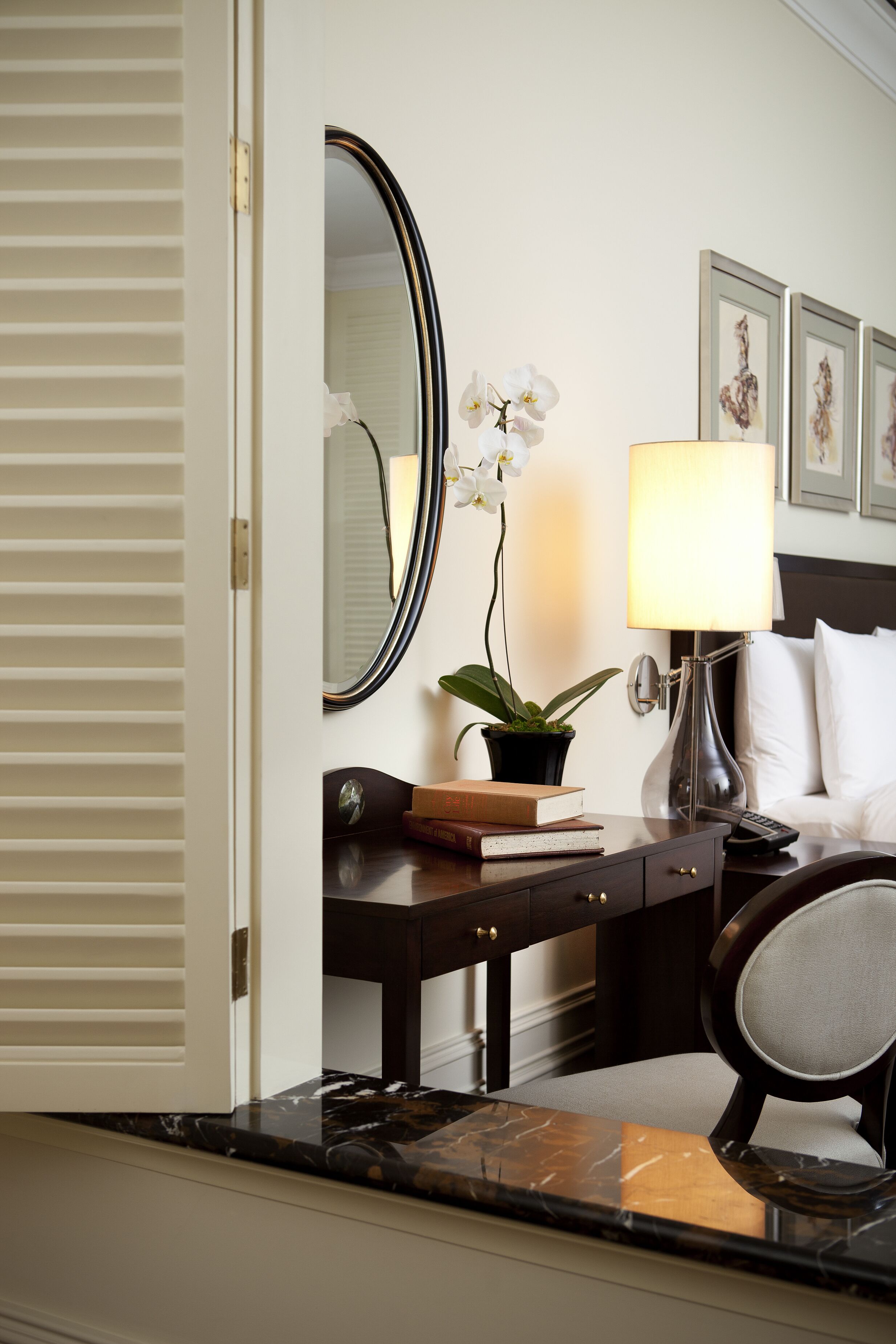 Een hotelkamer met een bed, een nachtkastje met een lamp, een spiegel, een potplant en kunstwerken in een lijst aan de muur.