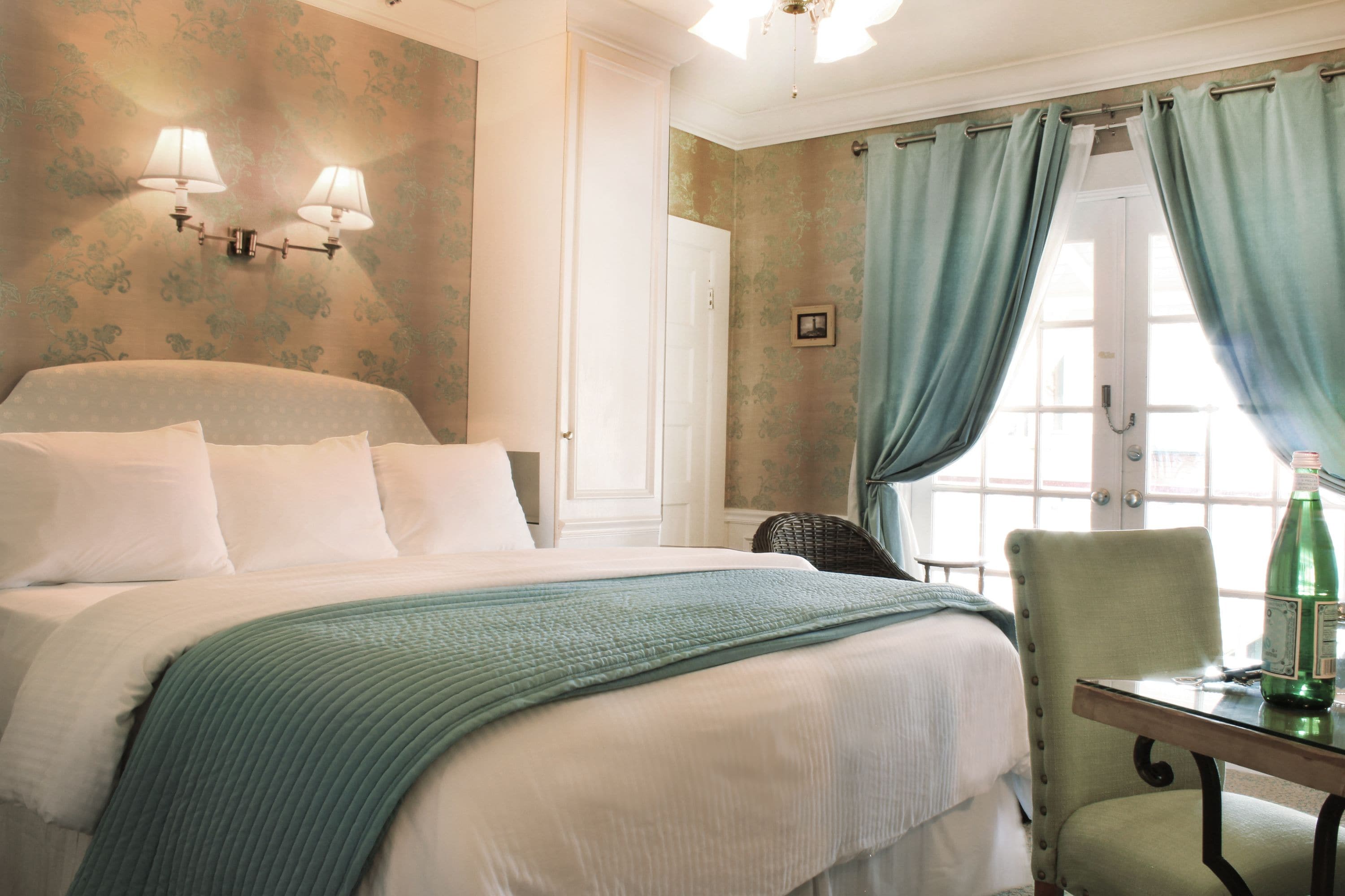 A hotel room with a large bed, a chair, a table with a bottle, and a window with curtains.