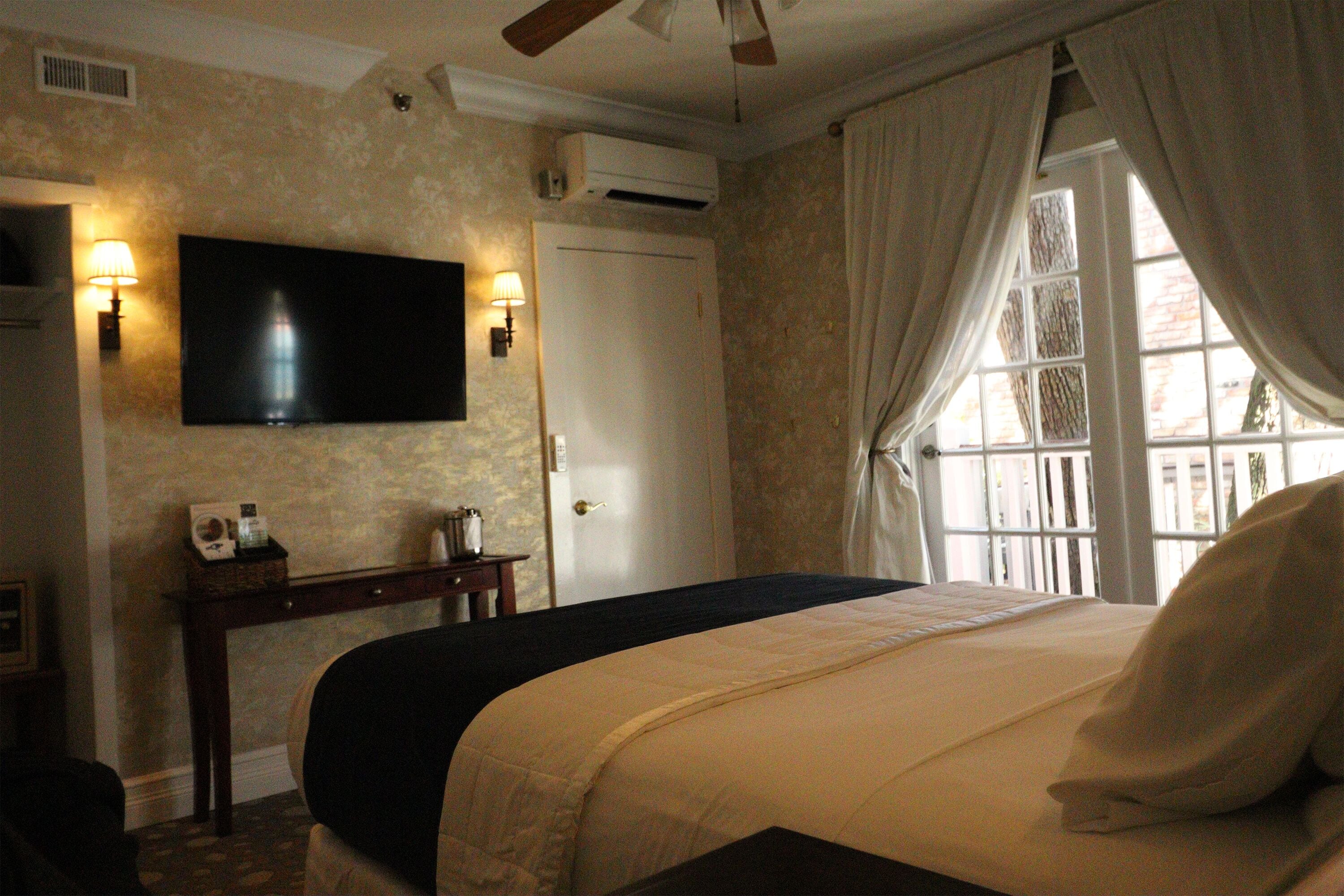 Quarto de hotel com uma cama, televisão de tela plana na parede, mesa de cabeceira com abajur e uma janela com cortinas.
