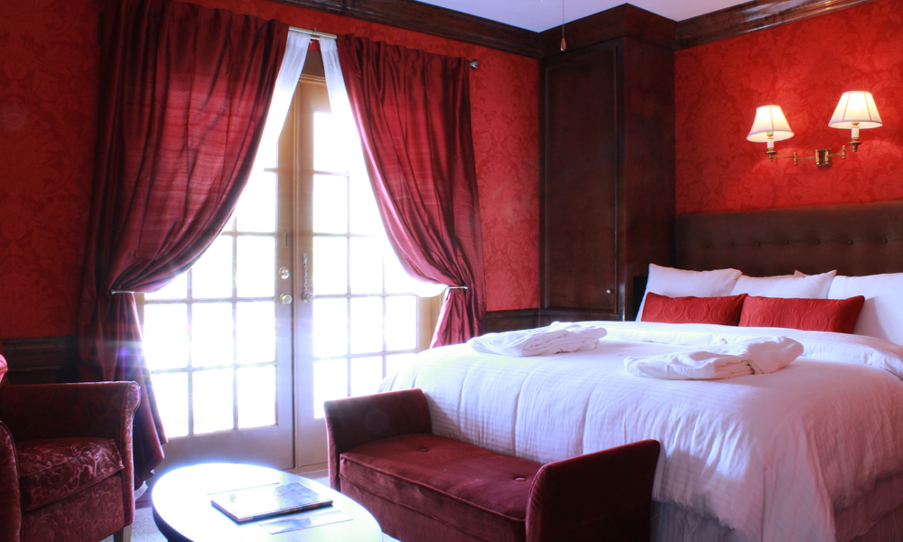 Quarto de hotel com uma cama grande, cortinas vermelhas, uma mesa redonda e duas cadeiras.