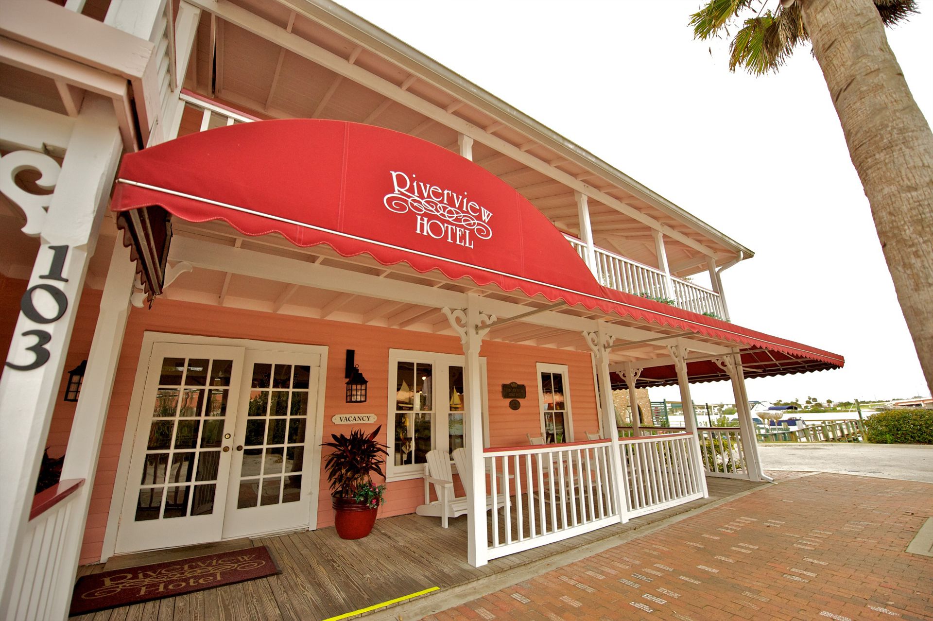 A two-story building with a red awning labeled "Riverwood Hotel." There is a "VACANCY" sign on the door, and a potted plant is visible outside.