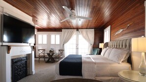 A room with a bed, a TV, a ceiling fan, a table with a lamp, and a fireplace. - The Riverview Hotel (New Smyrna Beach)