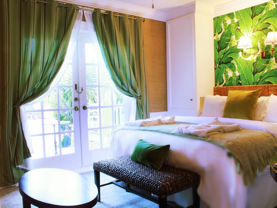A hotel room with a bed, a bench, a table, and a large window with curtains.
