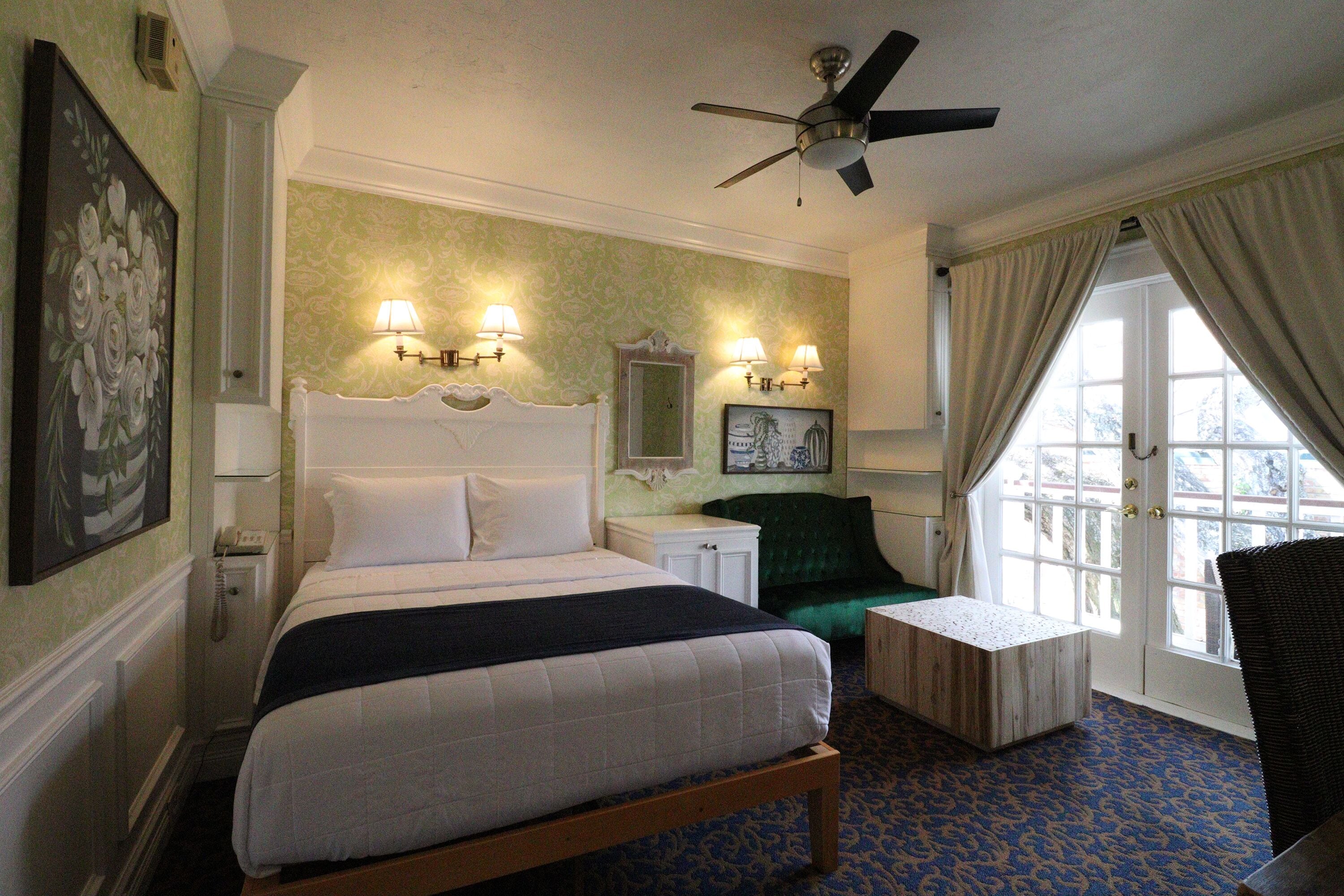 A hotel room with a large bed, two bedside tables, a chair, a desk, a ceiling fan, and a window with curtains.