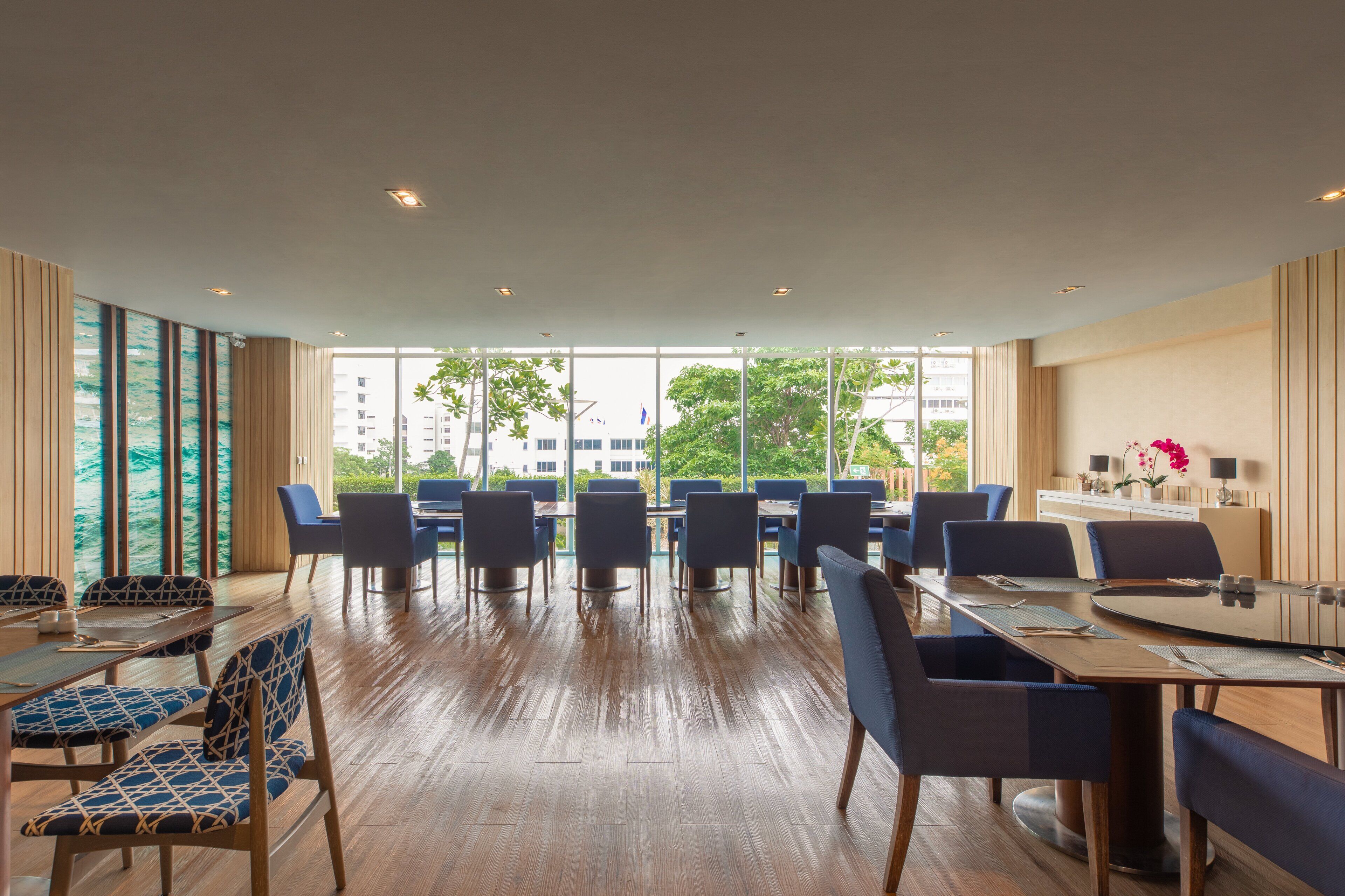 A modern conference room with a large glass table, blue upholstered chairs, and a view of buildings and trees through large windows.