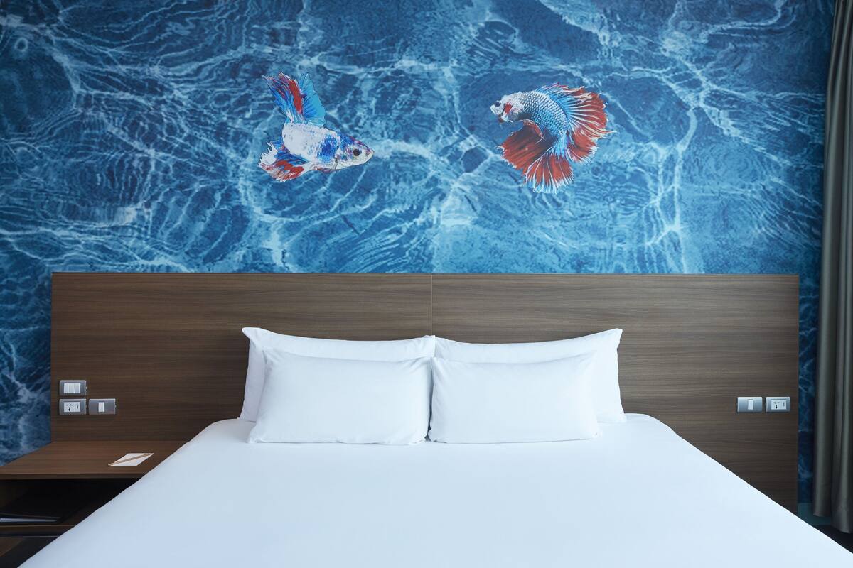 Habitación de hotel con cama, almohadas blancas, cabecera de madera y un mural en la pared con dos peces.