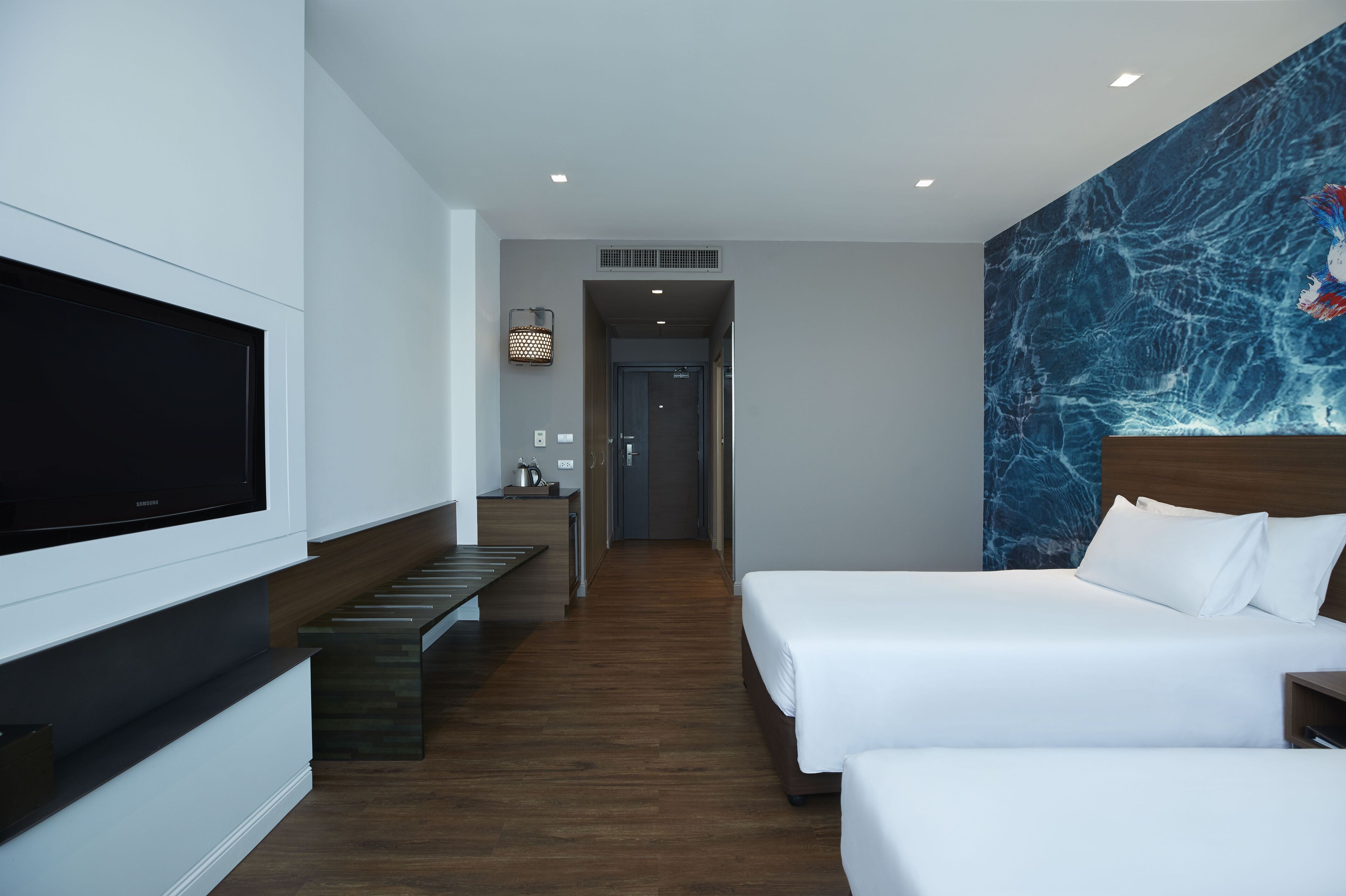 A hotel room with a large flat-screen TV, a wooden bench, two beds, a marble wall, and a hallway leading to a bathroom.