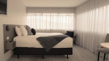 A modern hotel room with a large bed, a bedside table, a chair, and a view of the outdoors through sheer curtains.
