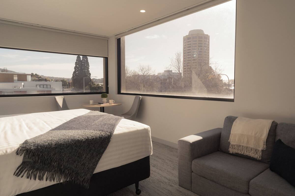 Quarto de hotel moderno com cama, uma poltrona cinza e uma pequena mesa.