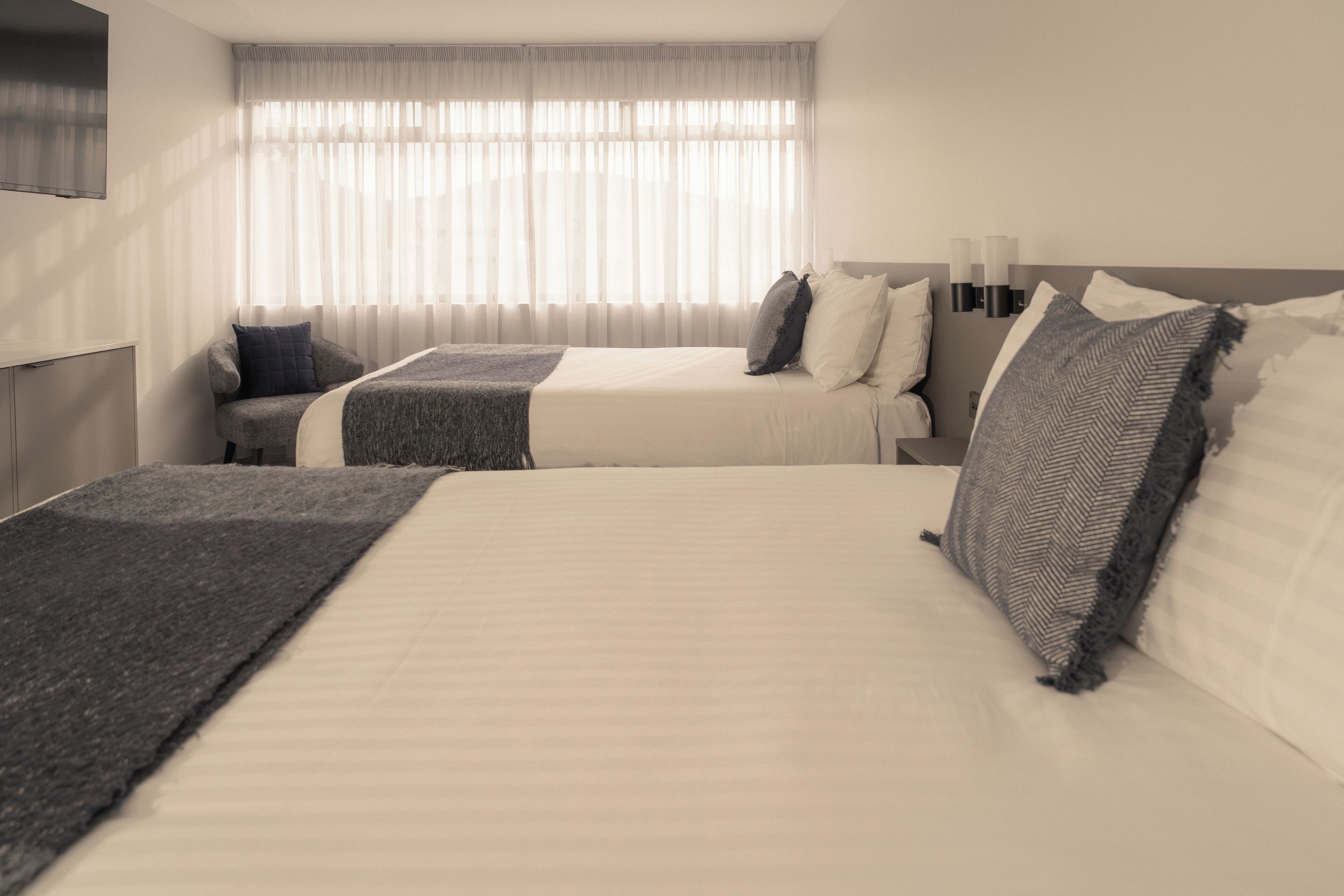 A hotel room with two beds, a large window with sheer curtains, a flat-screen TV mounted on the wall, and a bedside table with a lamp.