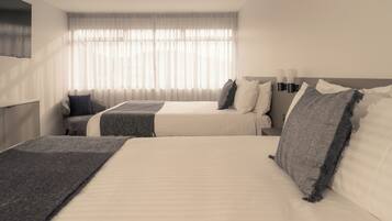 A hotel room with two beds, a large window with sheer curtains, a flat-screen TV mounted on the wall, and a bedside table with a lamp.
