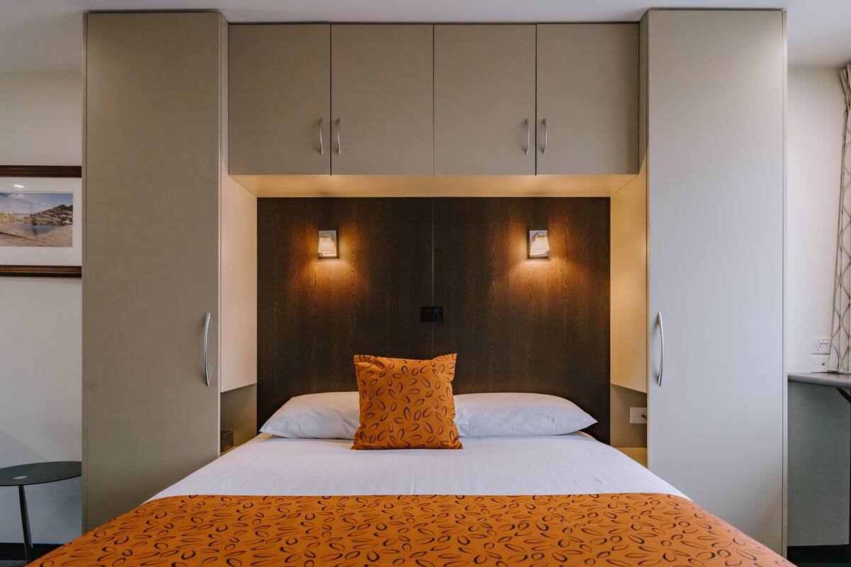 Quarto de hotel com uma cama, mesa de cabeceira, armário e duas luzes fixadas na parede.