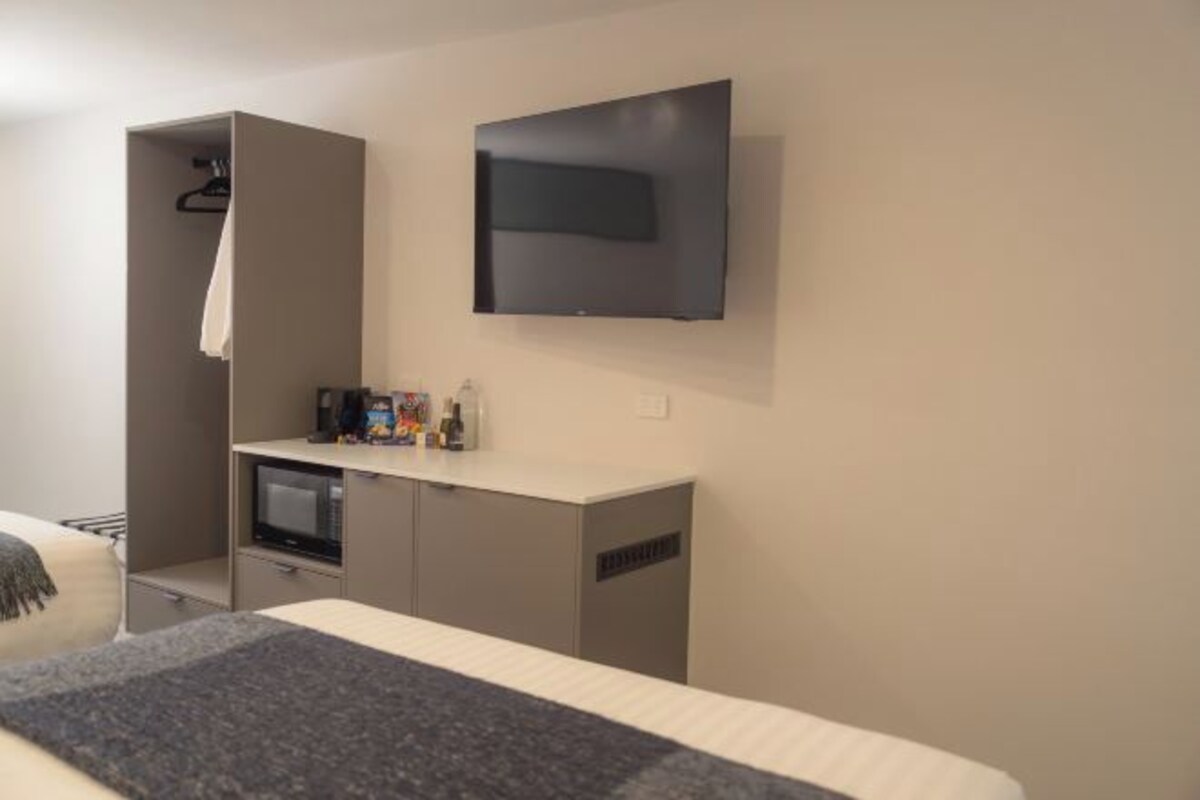 A hotel room with a bed, a TV mounted on the wall, a small kitchenette, and a wardrobe.