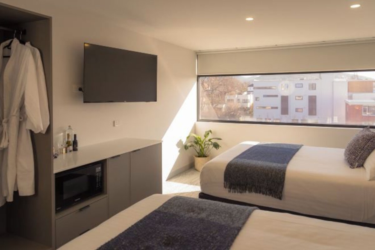 A modern bedroom with a TV mounted on the wall, a window with a view of buildings, a bedside table with a plant, and a bed with a blanket.