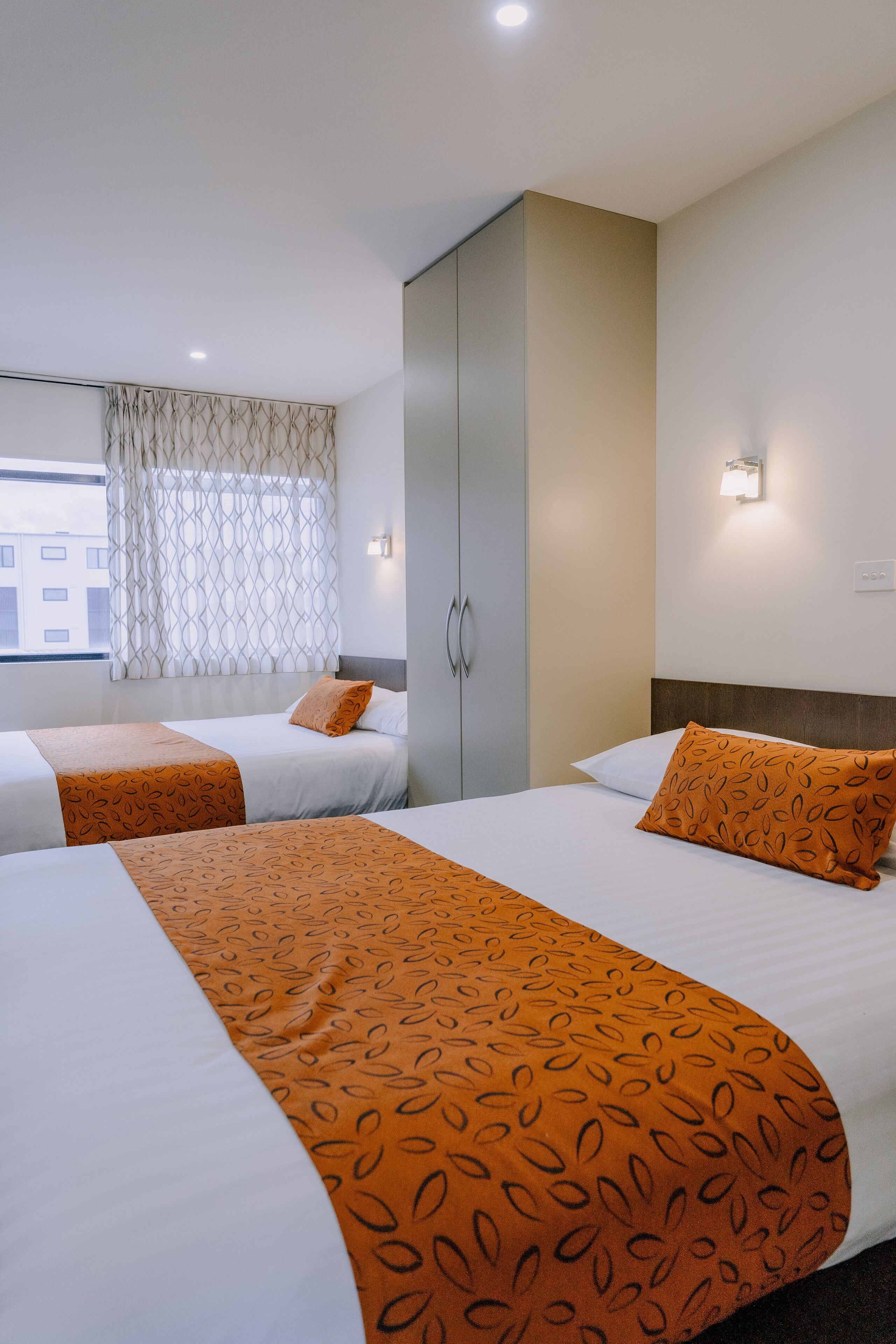 Habitación de hotel con dos camas, un armario, una ventana con cortinas y una lámpara fijada a la pared.