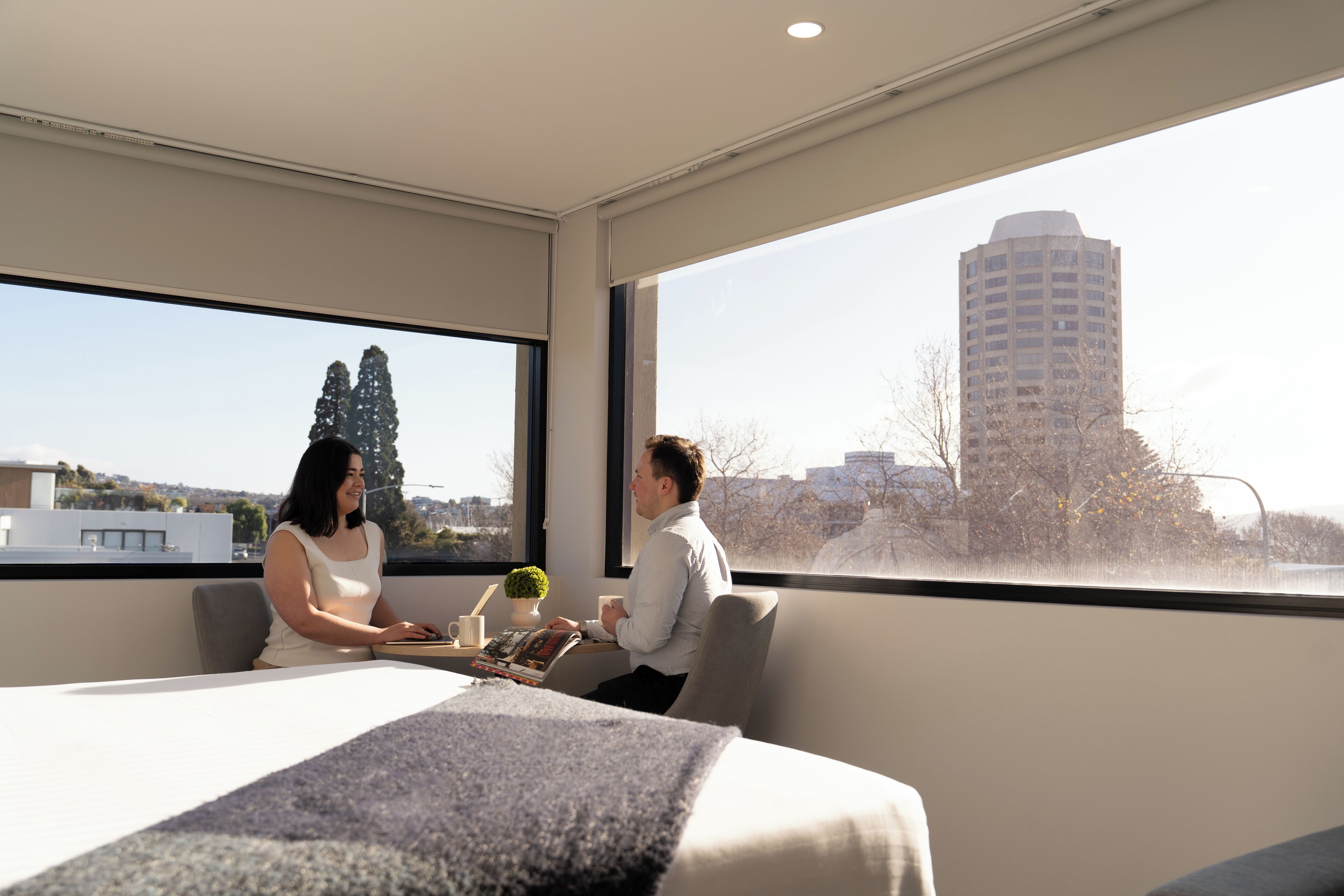 A modern hotel room with a large window, a bed, a table with a coffee cup, and a magazine.