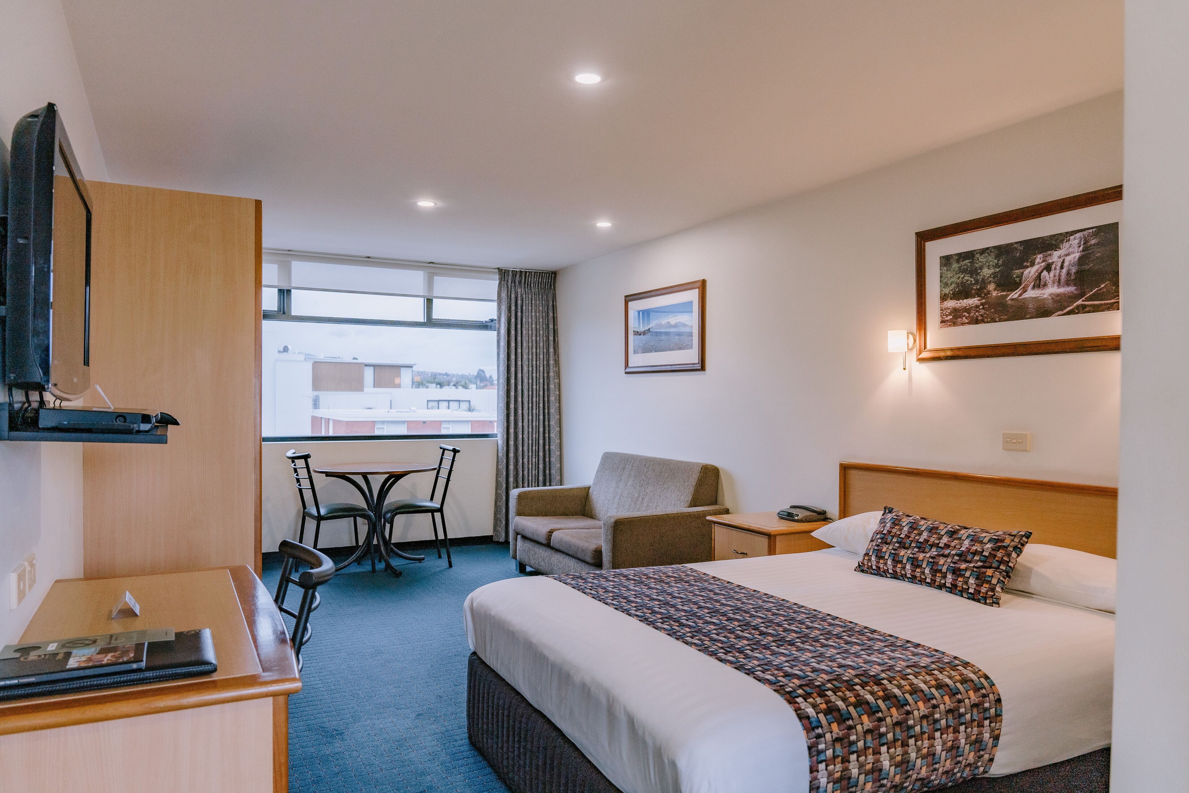 Habitación de hotel con cama, escritorio, silla, mesita y ventana con vistas a edificios.