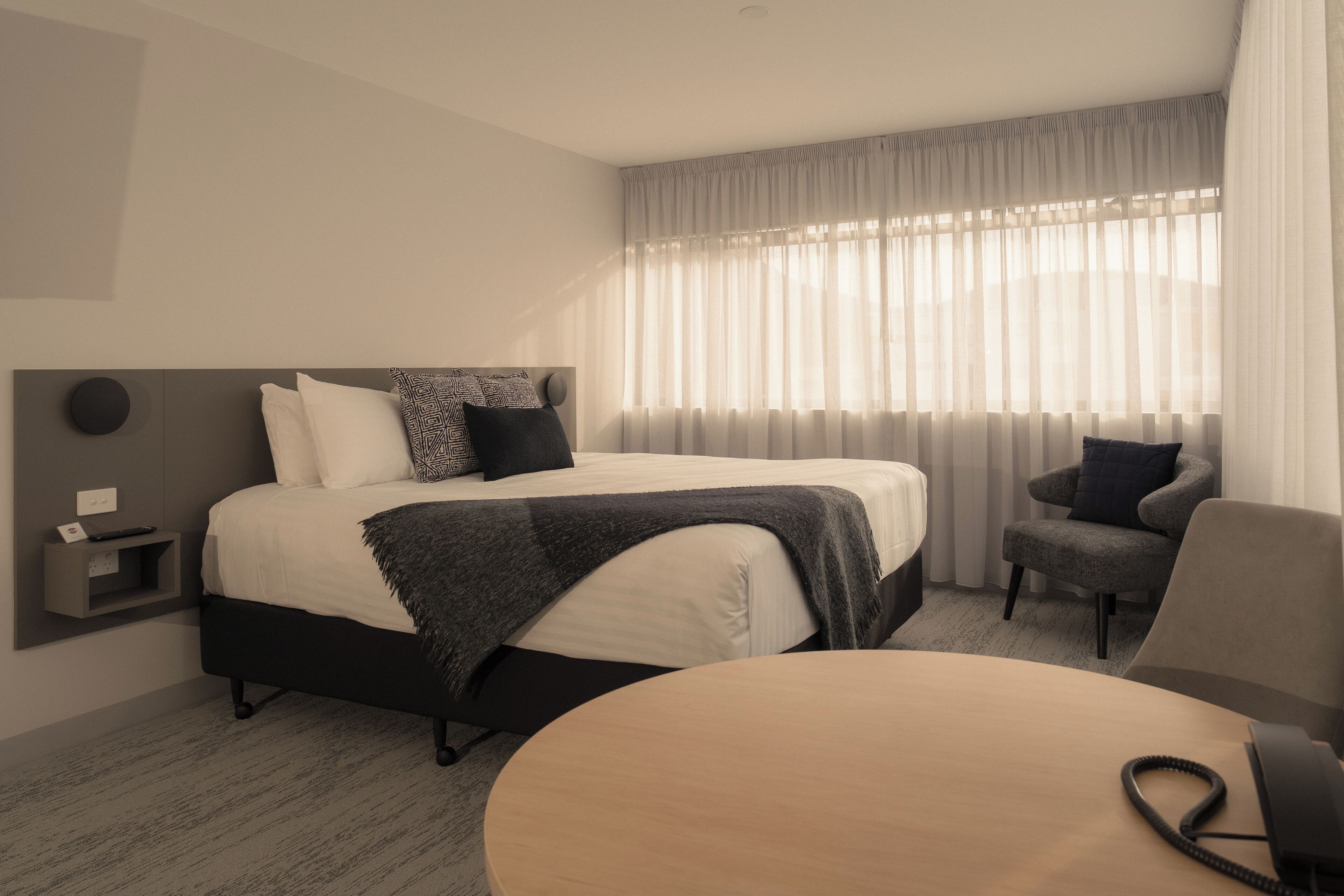 Una camera d'albergo moderna con un letto rifatto, un tavolo rotondo in legno, una sedia e un comodino con una lampada.