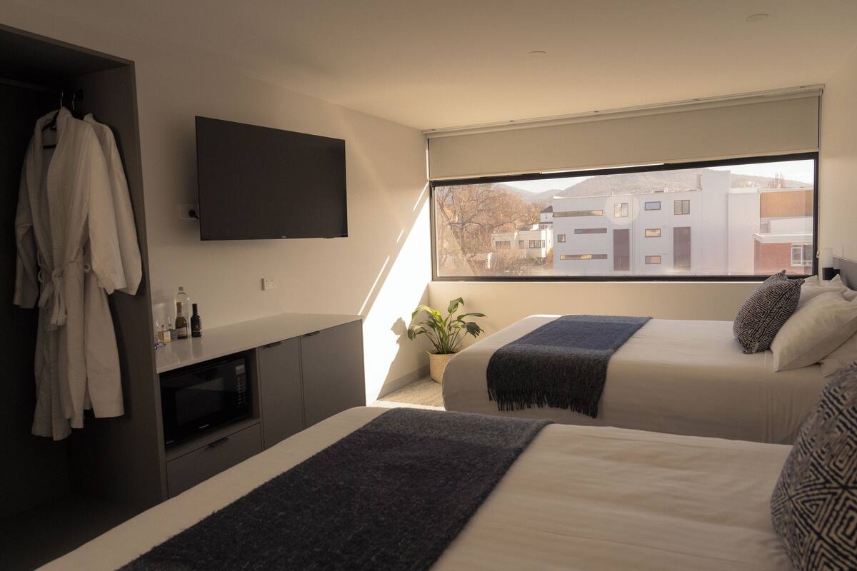 Quarto de hotel moderno com duas camas, televisão de tela plana, micro-ondas e uma grande janela com vista para edifícios e montanhas.