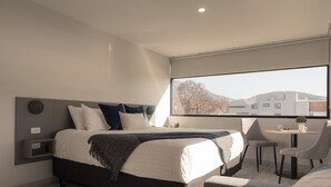 Un dormitorio moderno con una cama grande, un escritorio y vistas a edificios y árboles a través de una ventana.