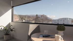 Una habitación con vistas a montañas, una planta en maceta, una silla, una mesa con un libro y una taza, y un jarrón con una planta.