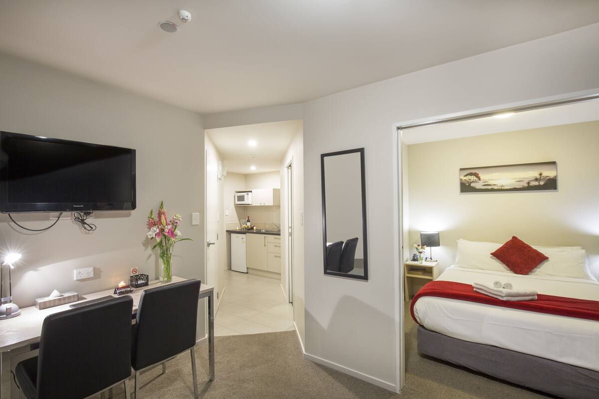Ein Hotelzimmer mit einem Bett, einem Schreibtisch mit Stühlen, einem Fernseher, einem Bild und einer Küche im Hintergrund.