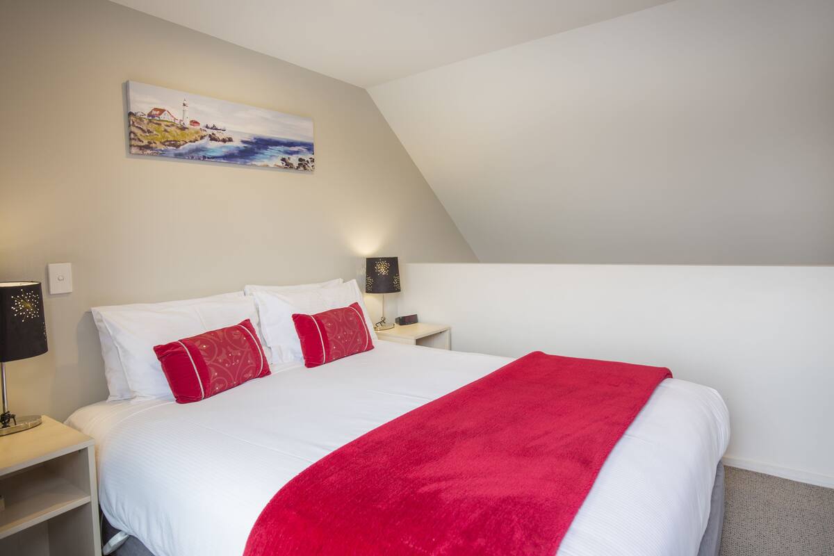 Ein Hotelzimmer mit Bett, weißer Bettwäsche, roten Kissen, einer roten Tagesdecke, einem Bild an der Wand und einem Nachttisch mit Lampe.