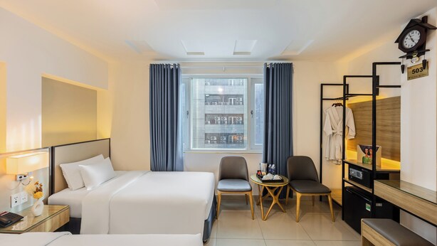A hotel room with two beds, a small table with chairs, a desk with a lamp, a clock, and a view of the city through a window.