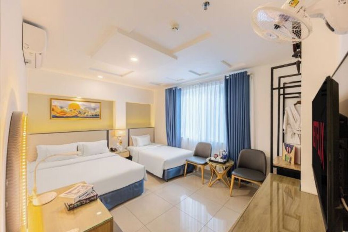 A hotel room with two beds, a desk, a chair, a small table, a TV, and a window with curtains.