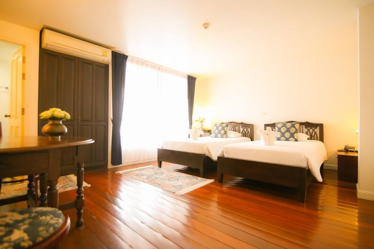 Une chambre avec deux lits, un plancher en bois, une grande fenêtre avec des rideaux, une petite table avec un vase et une table de chevet avec une lampe.
