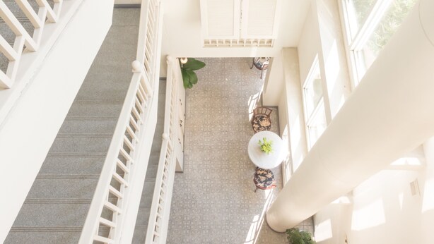 Un escalier à haute voûte, doté de rampes et de volets blancs, mène à un balcon dont le sol est orné de motifs et où se trouve une petite table avec une plante.