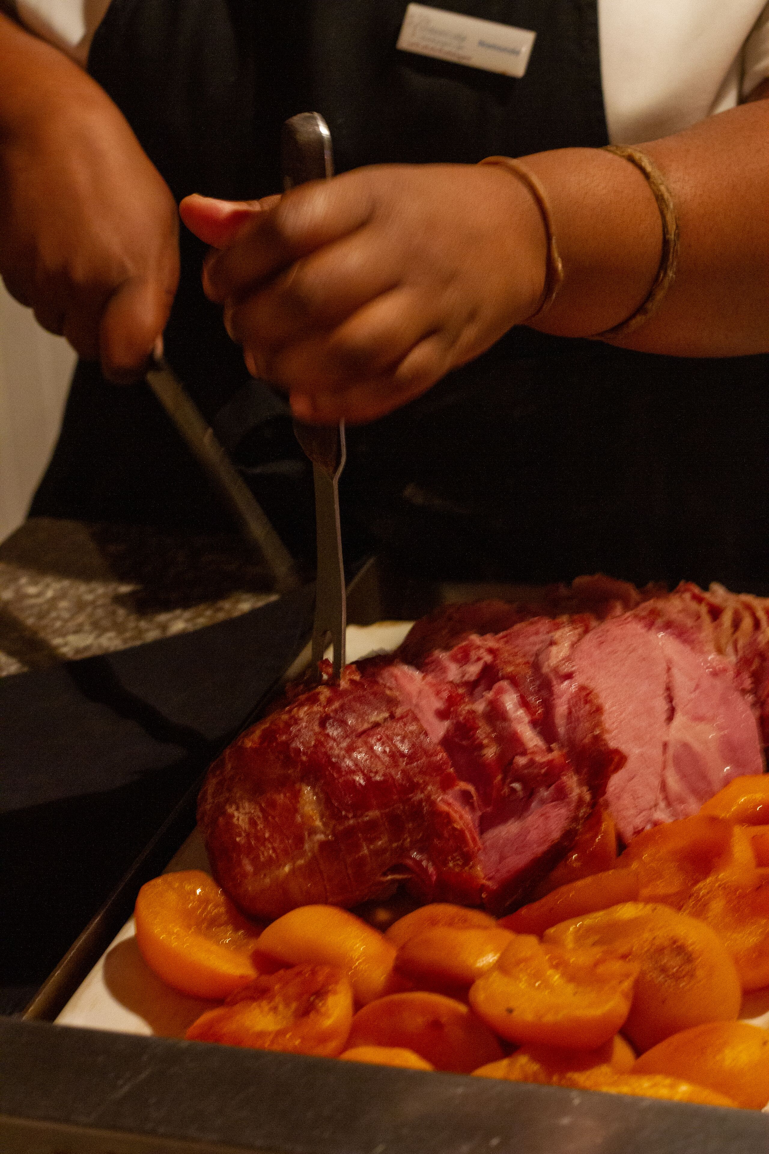 A person slicing a large piece of meat, likely ham, on a cutting board with sliced oranges nearby.