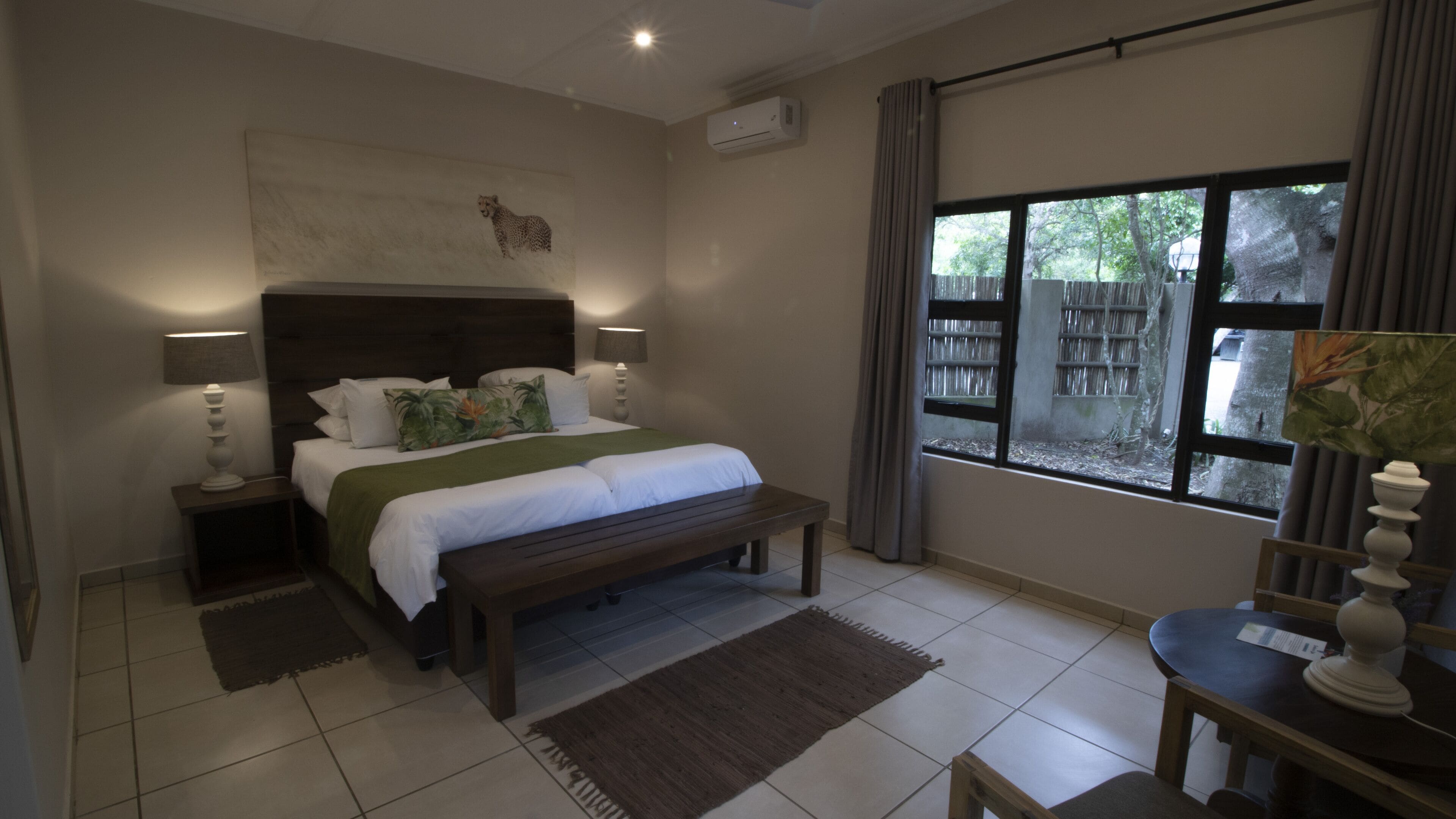 A bedroom with a large bed, bedside lamps, a bench, a desk, and a window with a view of trees.