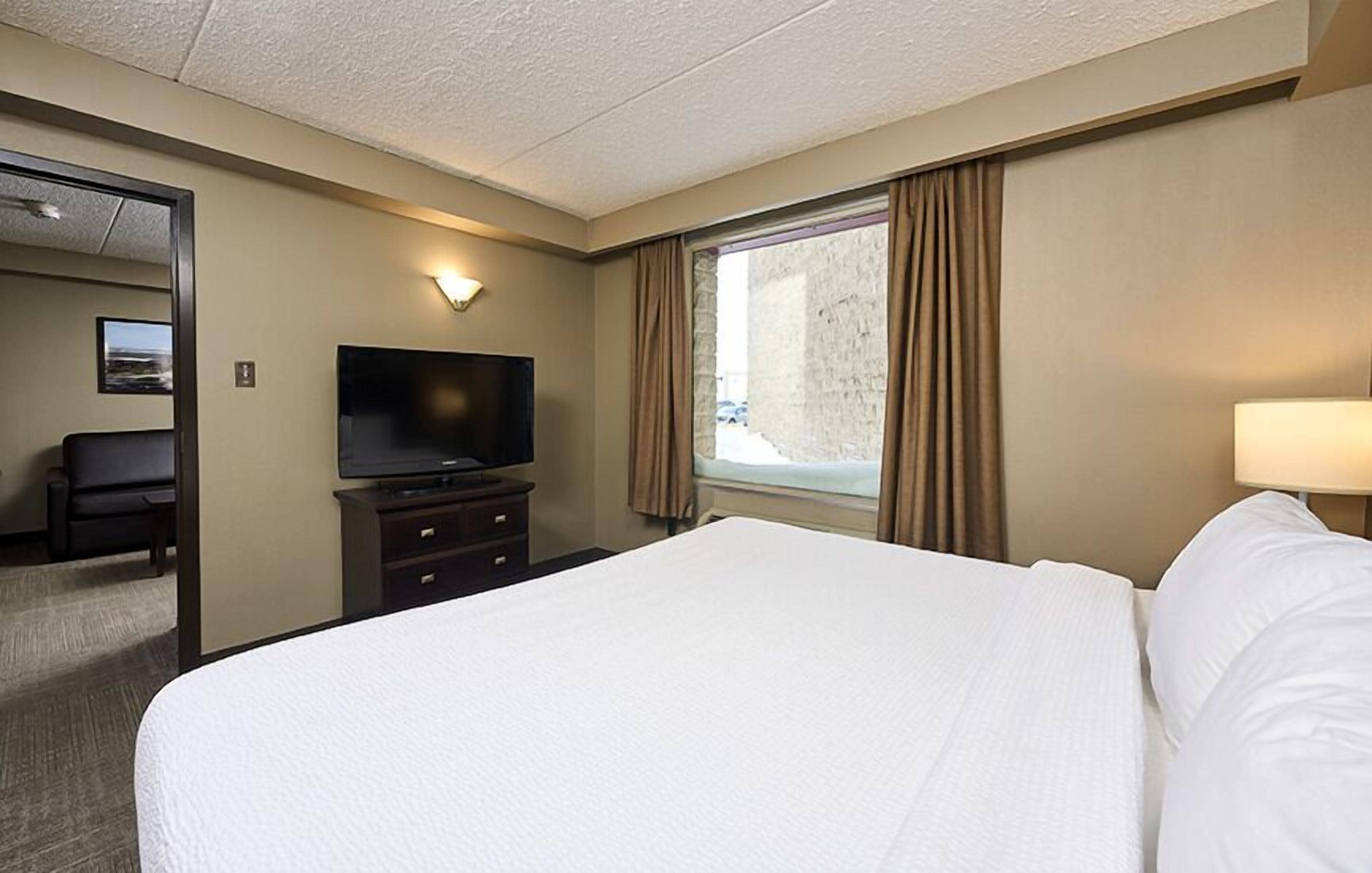 A hotel room with a large bed, a flat-screen TV on a dresser, a window with curtains, and a mirror in the corner.