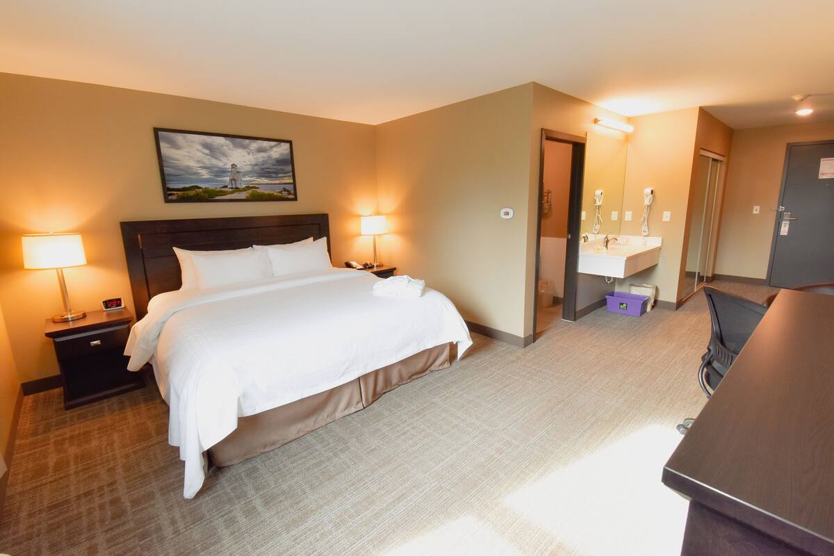 Habitación de hotel con cama, lámparas de noche, escritorio, espejo y baño con lavamanos y toallero.