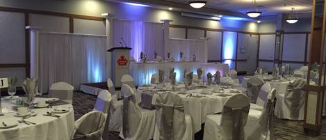 Un salón de banquetes con mesas redondas dispuestas para un evento formal, manteles blancos y sillas con cintas grises.