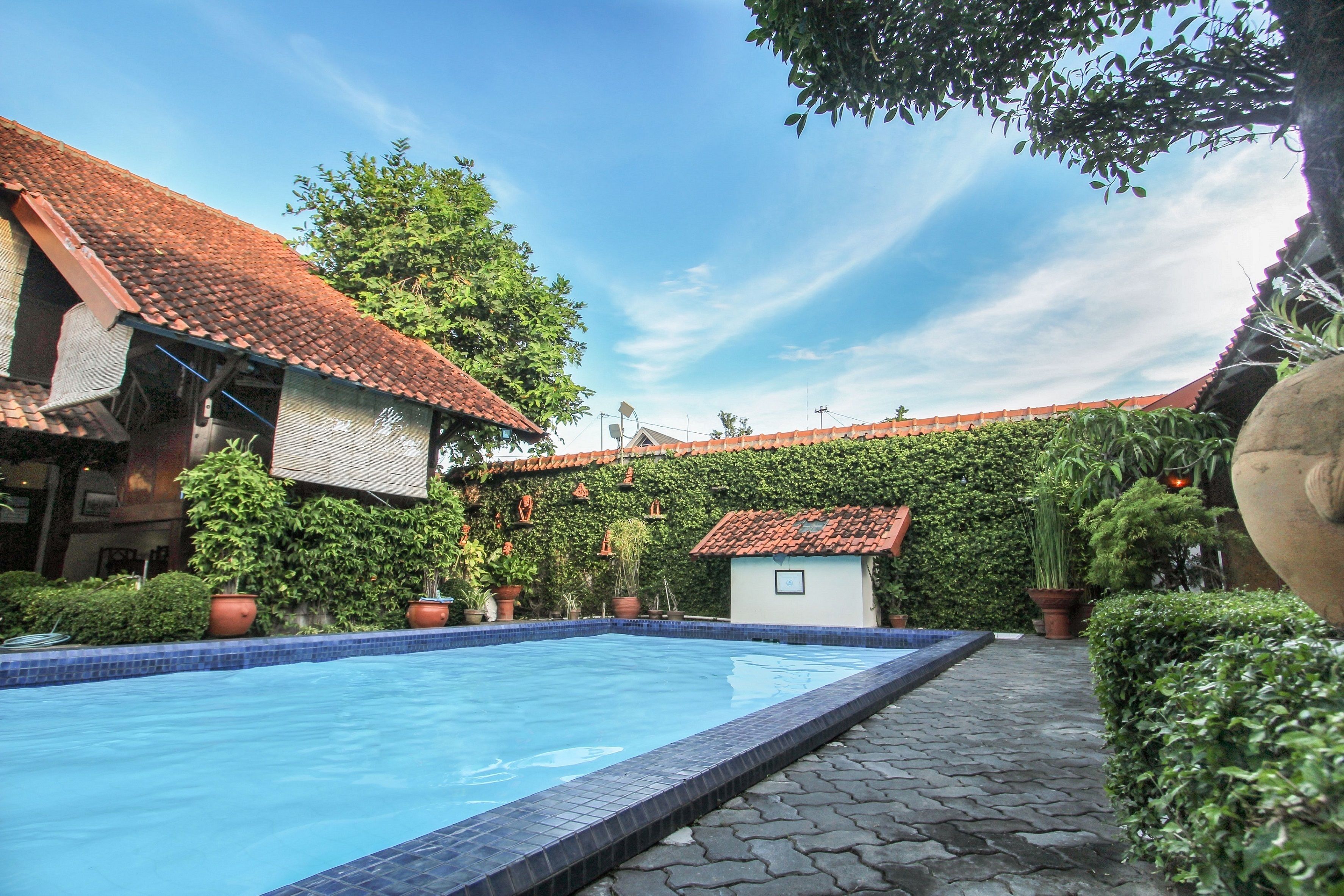 A swimming pool with a tiled edge, surrounded by potted plants and a building with a tiled roof.