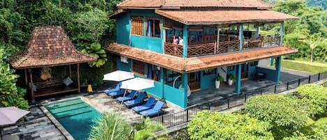 Uma casa de dois andares com piscina, varanda coberta e vegetação exuberante.