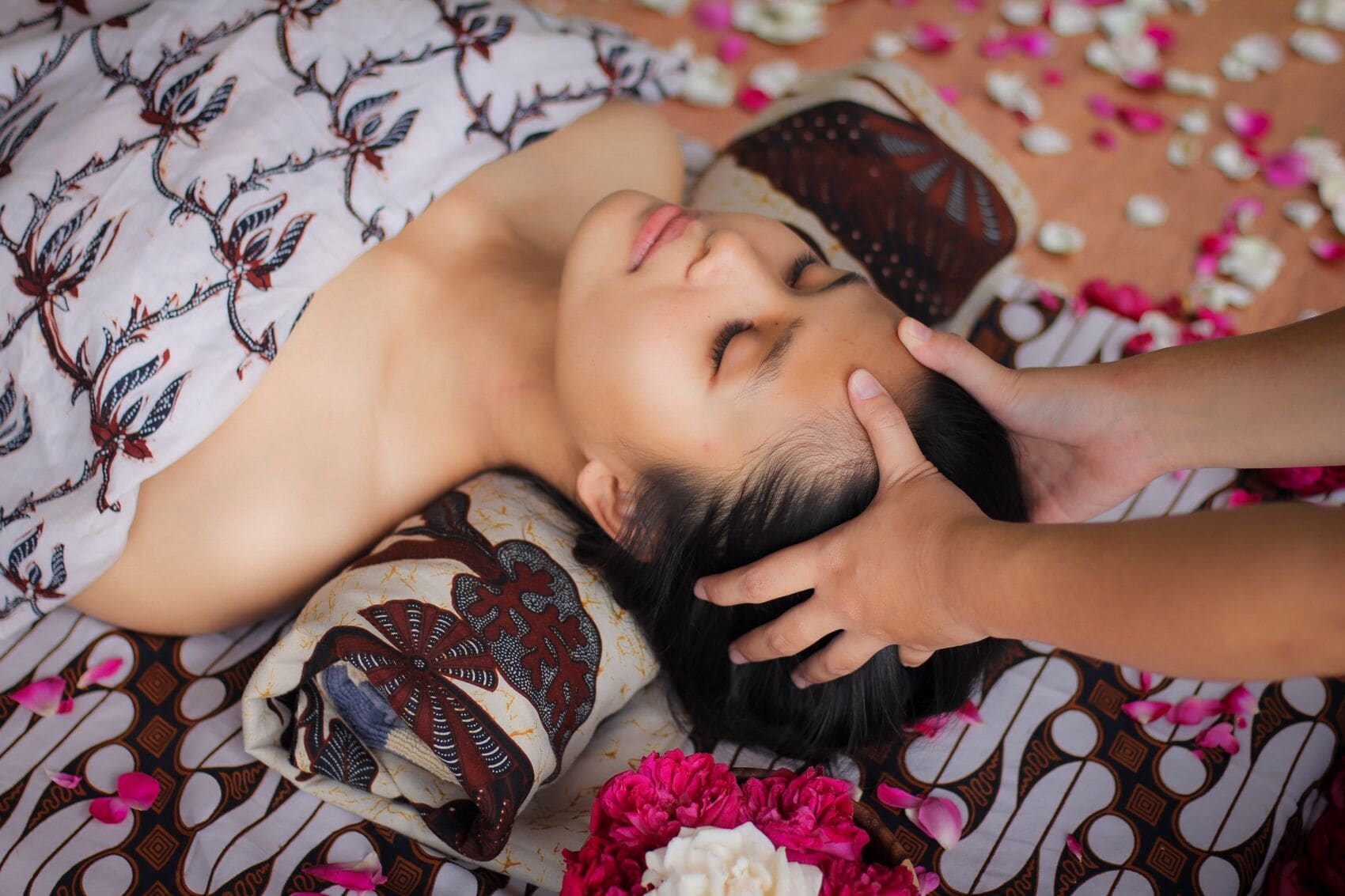 Une personne allongée sur un lit, recouverte d’une literie à motifs, avec des pétales de fleurs éparpillés autour.