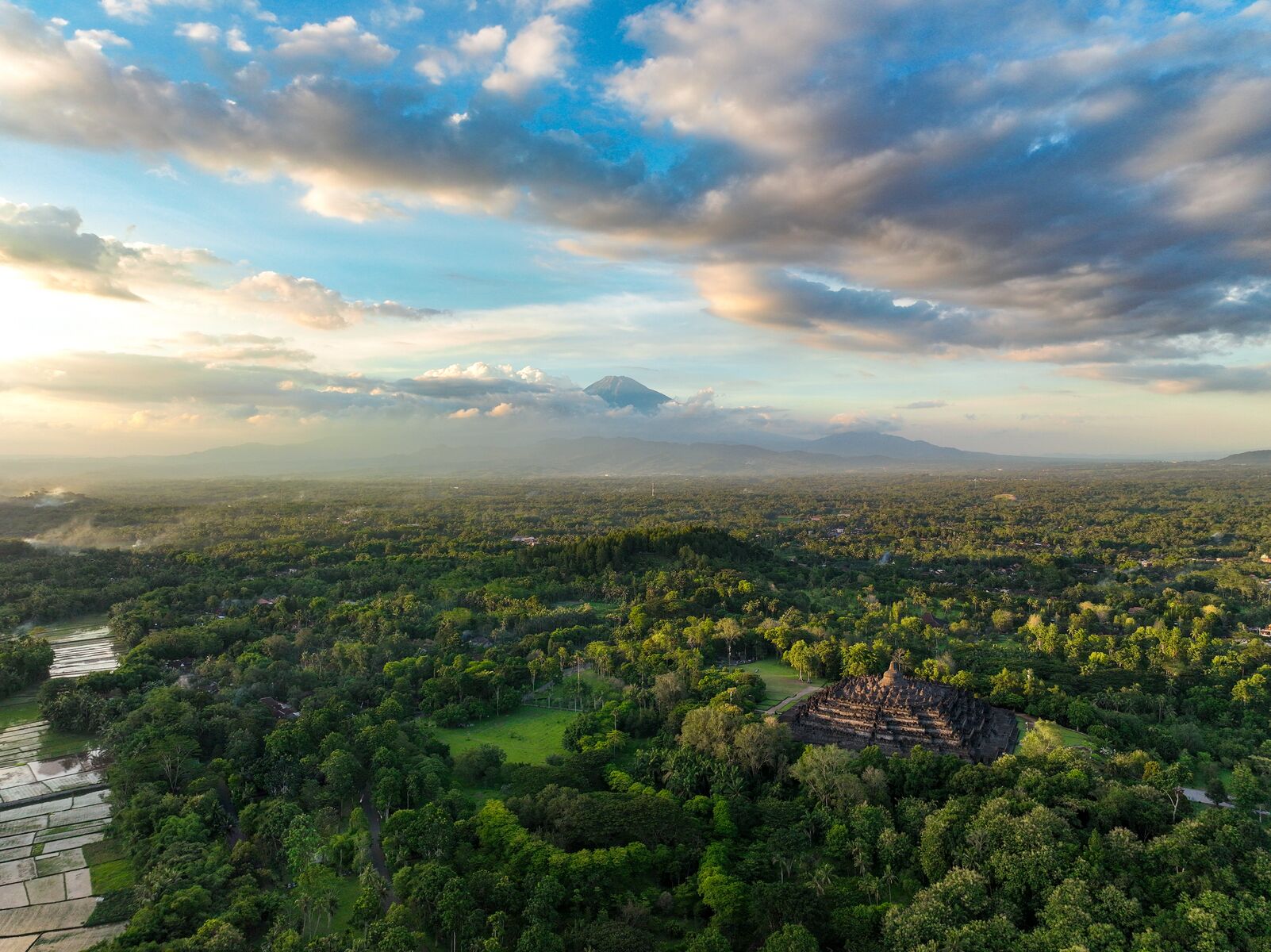 Vista aérea de un exuberante paisaje con una imponente pirámide antigua rodeada de vegetación densa y una cordillera distante.