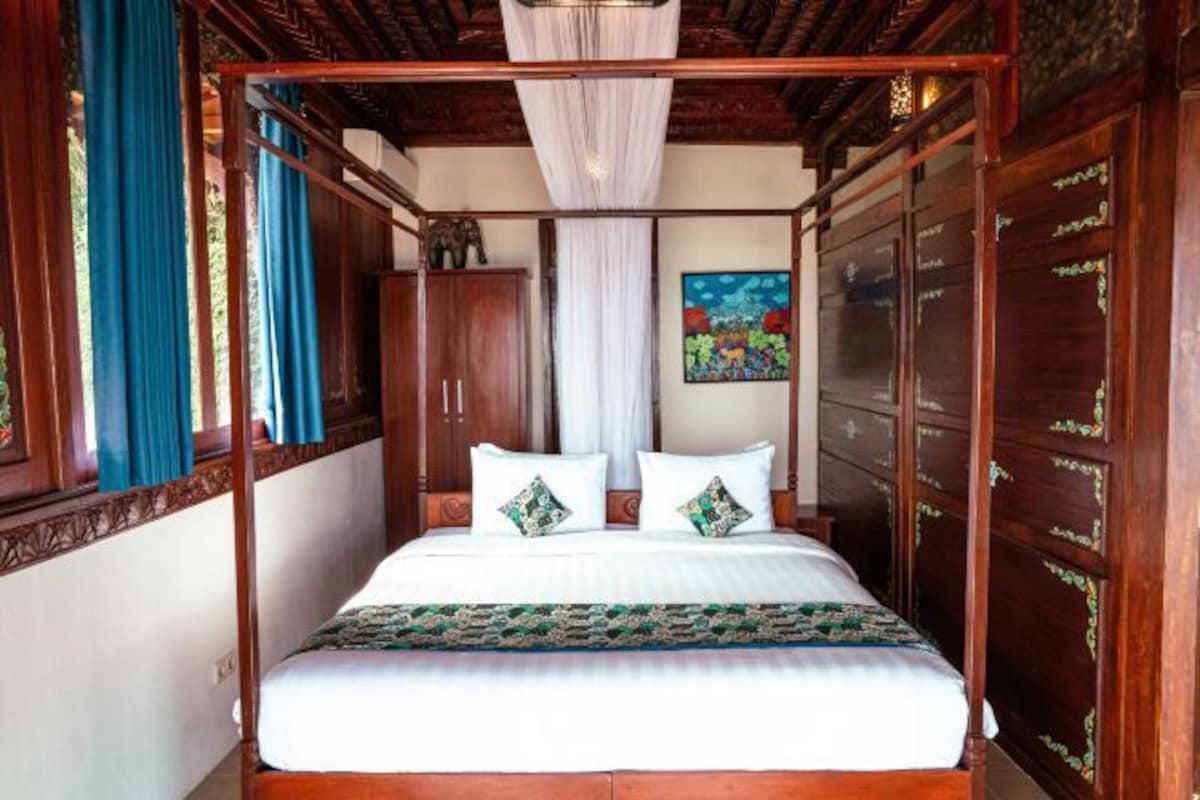 A four-poster bed with white linens and decorative pillows, a wooden wardrobe, and a painting on the wall.