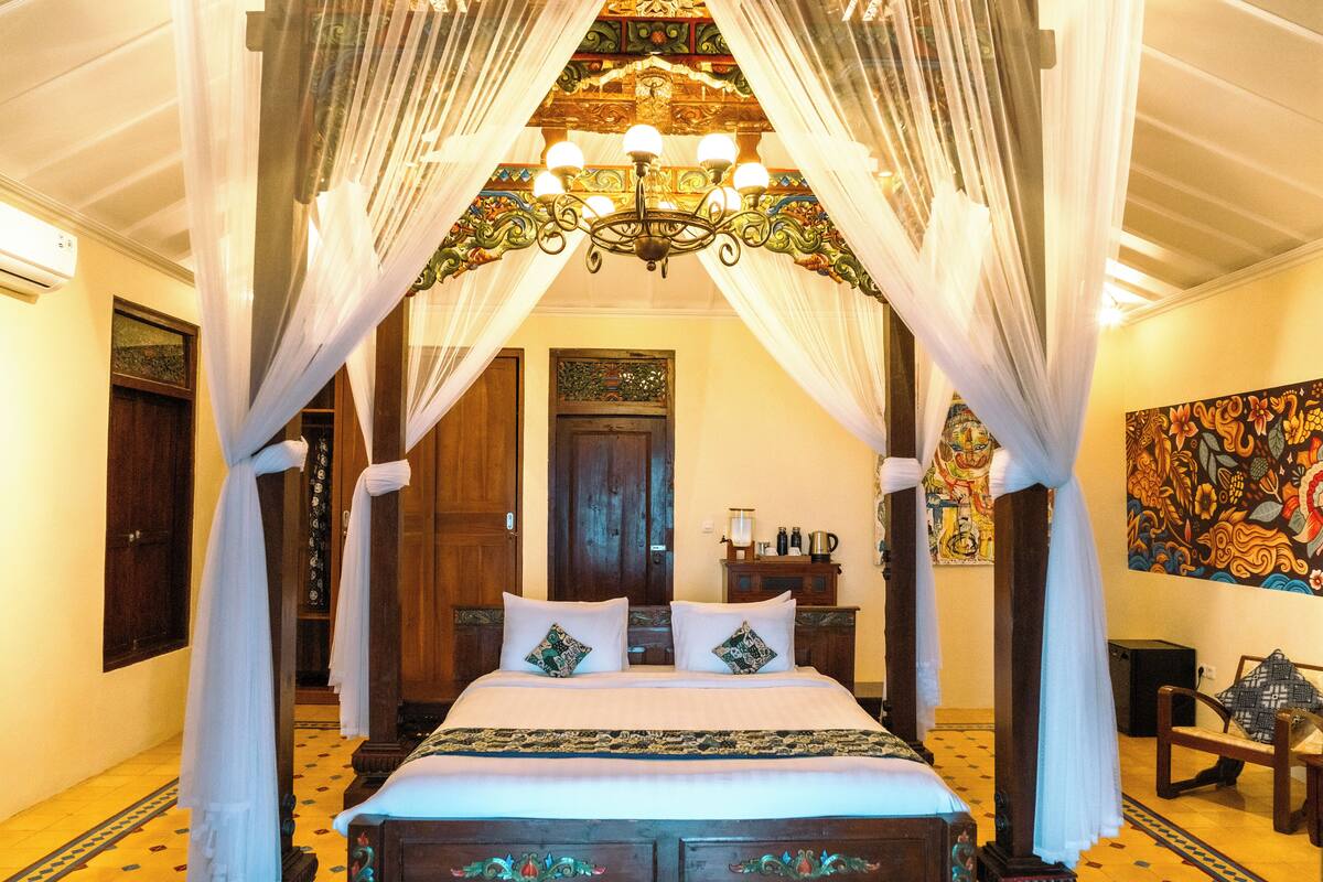 A four-poster canopy bed with a chandelier, a wooden door, and a colorful tapestry on the wall.