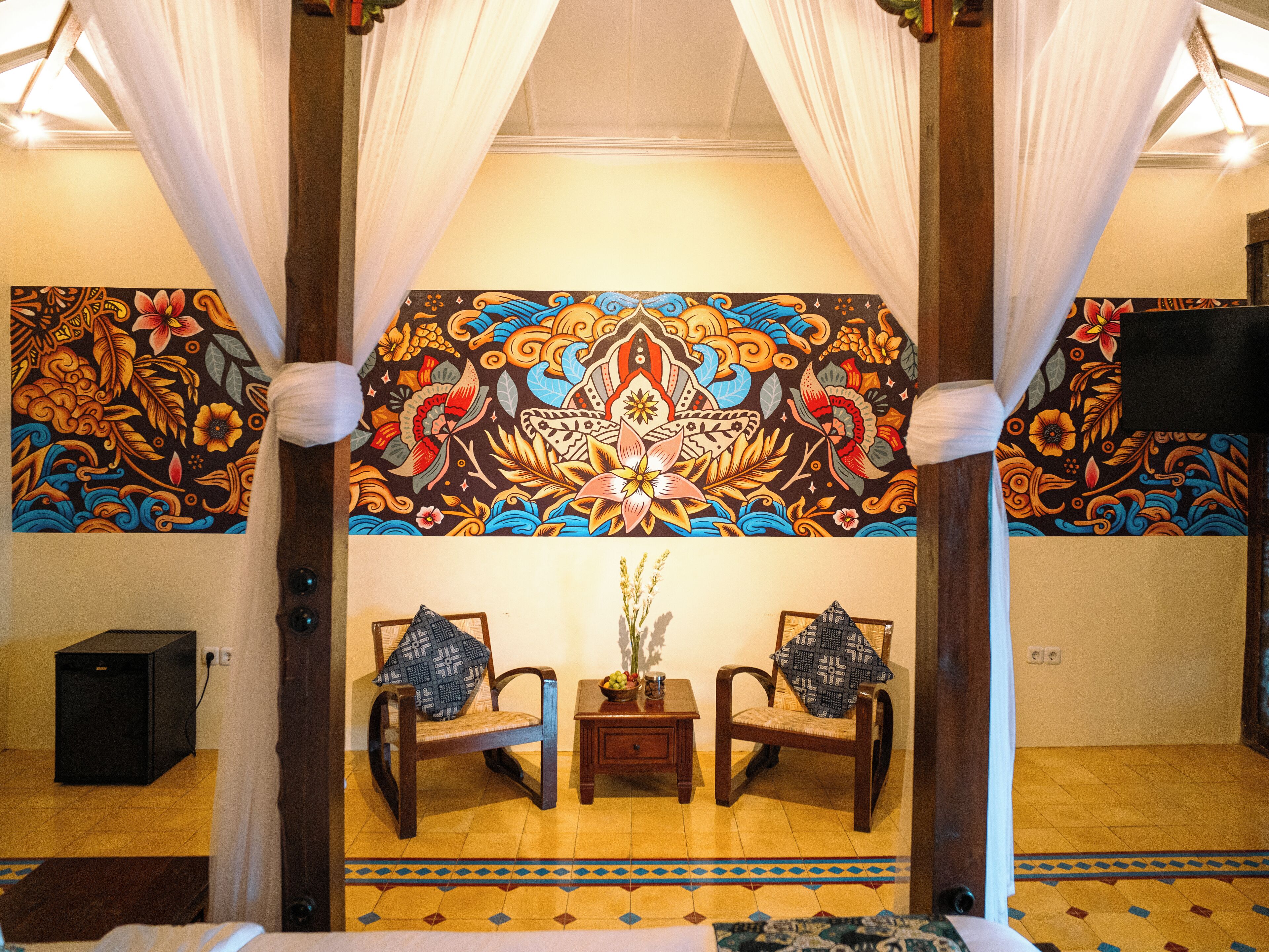 A room with a traditional design, featuring a bed, two chairs, a small table, and a large decorative tapestry on the wall.
